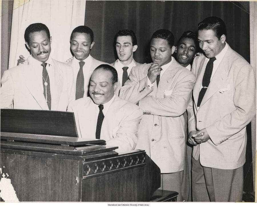 8 x 10 inch photograph; Count Basie playing the piano observed by six unidentified men, one of whom is holding drum sticks