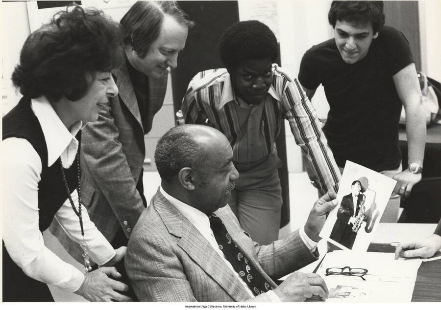 8 x 10 inch photograph. From left to right: Paula Berger (mother of photographer Ed Berger); Dan Morgenstern; Vincent Pelote; David Penque. Benny Carter is sitting in the middle of the group holding up a photograph of an unidentified saxophonist.