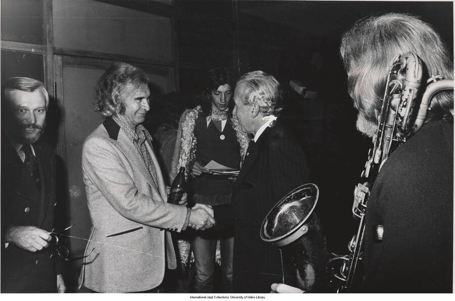 8 x 10 inch photograph; Dave Brubeck shaking hands with an unidentified man in the presence of other persons including baritone saxophonist Gerry Mulligan.