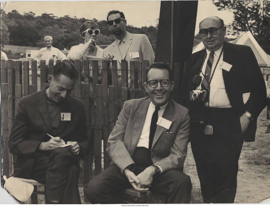 11 3/4 x 15 3/4 inch photograph; Leonard Feather with unidentified persons, including a photographer, wearing a "staff" name tag with the letter "J" on it