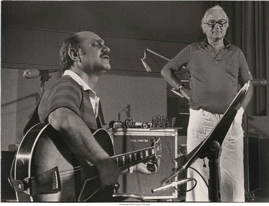 11 x 14 inch photograph; Joe Pass playing the guitar and Norman Granz standing by. In the background are Brazilian instruments on top of a box with the name: Paulinho da [Costa]