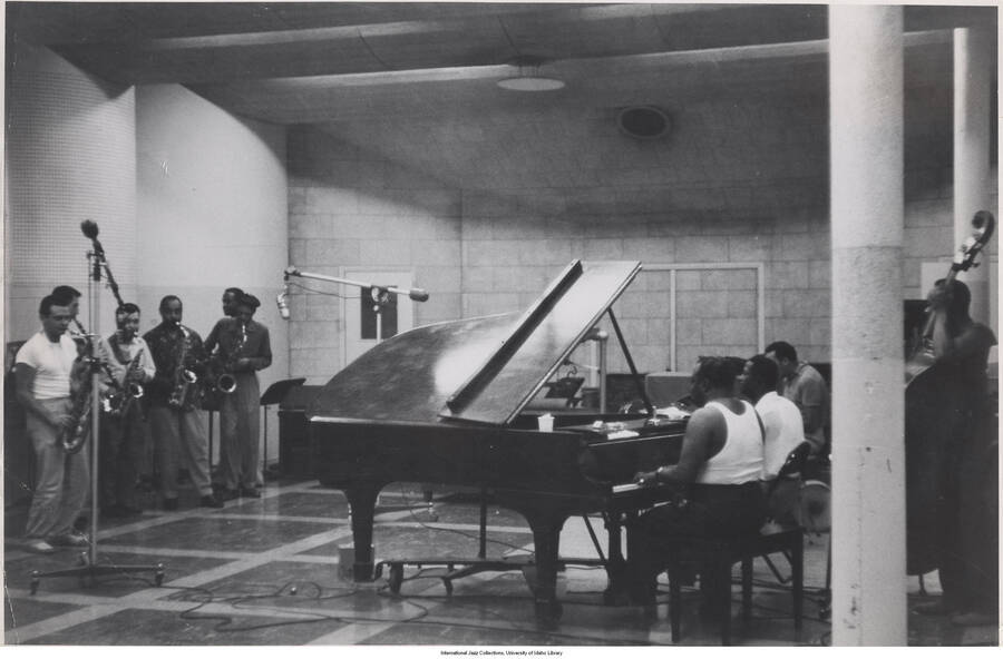 8 3/4 x 13 1/4 inch photograph; an unidentified band performing in a recording studio. Among the musicians present are Stan Getz, in white t-shirt at extreme left, Benny Carter, fourth from left, and Count Basie, at left on the piano bench.