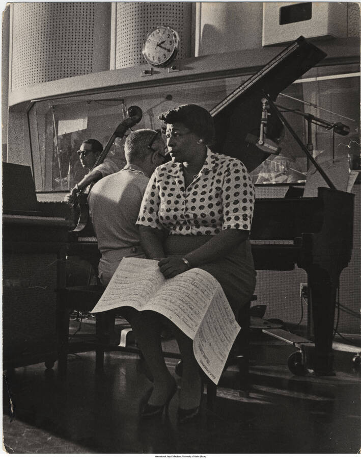 14 x 11 inch photograph; Ella Fitzgerald sitting on a piano bench beside an unidentified pianist (Jimmy Rowles?), with her back to the piano in a recording studio. She is holding a score of My Old Flame.  Also present is an unidentified man leaning on a bass violin. Handwritten on the back of the photograph: Donahue & Coe (Mort Nasatir)