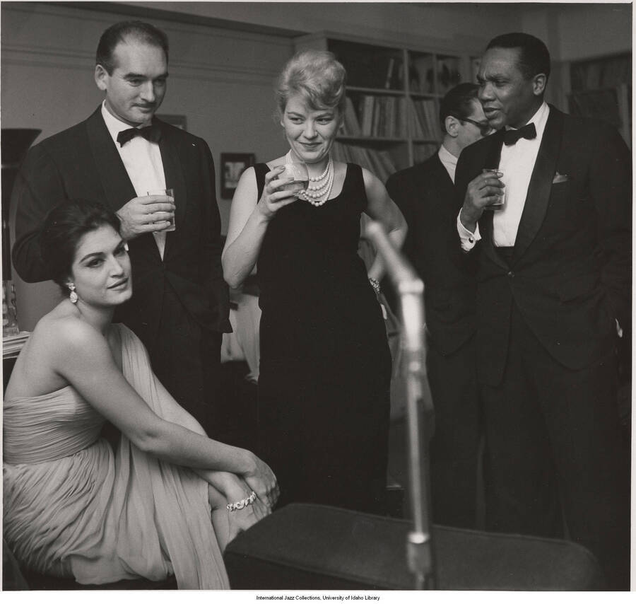 14 x 11 inch photograph; Jane and Leonard Feather with unidentified persons in a reception, probably in their apartment