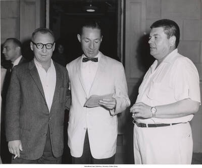 8 x 10 inch photograph; Leonard Feather reading from a program, beside Charles Dederich, founder of Synanon, and an unidentified man