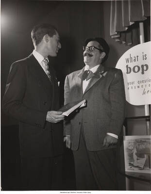 10 x 8 inch photograph. Label on the back of the photograph reads: At Bop City, Leonard Feather (left) offers his new book, "Inside Bebop," to Jerry Colonna as an answer to Jerry's questions about the new jazz. Note Colonna's bop beret, tie, and glasses