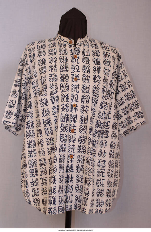 Blue/gray/white, cotton w/indigo calligraphic figures all over in rows and columns.  Indigo blue has migrated (bled) into white fabric.  Shirt tail style, short sleeves w/ standing band collar.  No labels, looks homemade.  6 brown wooden buttons center front placket.  R and L breast pockets.