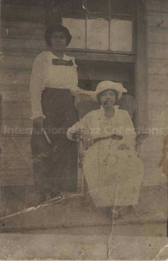 Two unidentified women posing on the porch of a house. One is standing up with her left arm resting on the back of a rocking chair in which the other is sitting