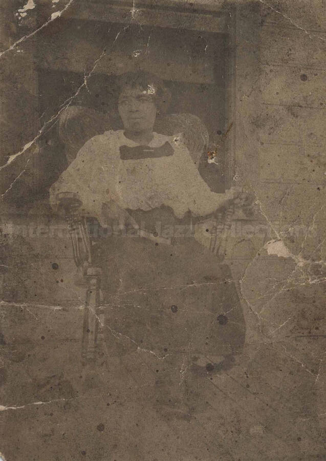 Unidentified woman sitting down in a rocking chair on the porch of a house (1 duplicate)