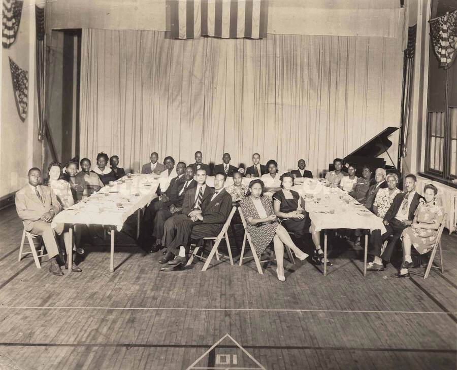 Unidentified persons sitting at a dinner table in what appears to be the gym or auditorium of a school. Stamped on the back of the photograph: Young Men's Christian Association, Pottstown, Pennsylvania