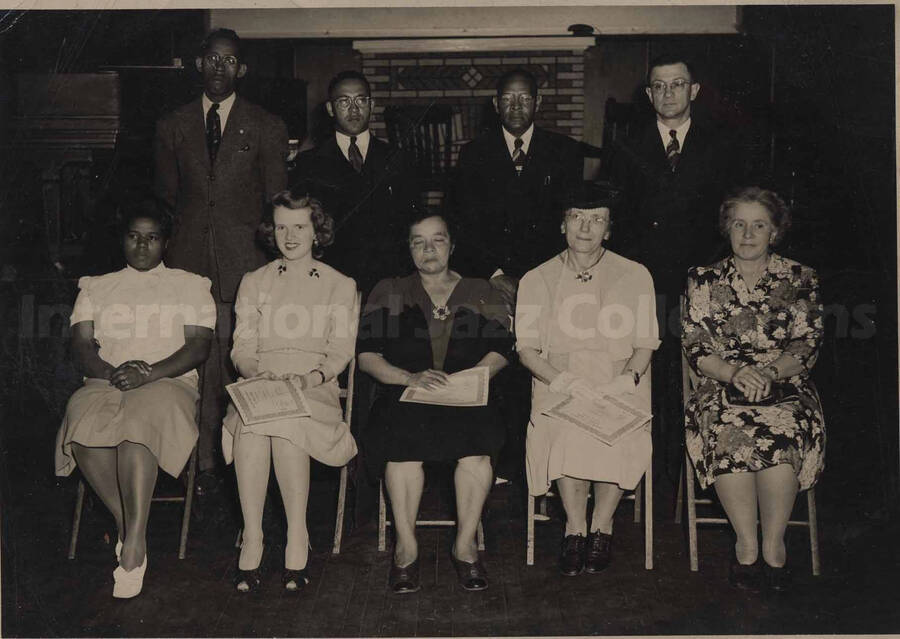 Four unidentified men standing behind five unidentified women who are sitting on chairs. Three of the women are holding certificates