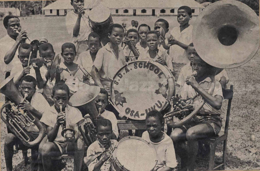 A band composed of boys. Written on the drum: S. D. A. School Osa-Uku. This is a clip from a magazine glued to a piece of cardboard