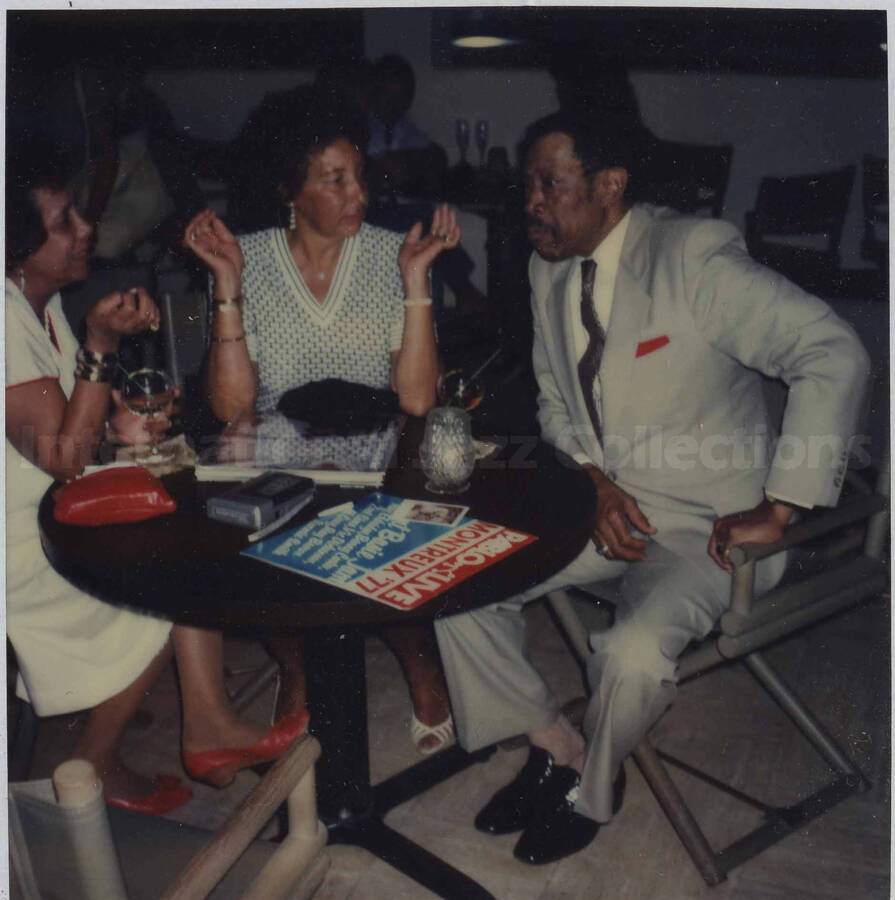 Al Grey sitting at a table in company of two unidentified women. On top of the table is the LP entitled "Pablo Live - Montreux '77 - Oscar Peterson Jam"