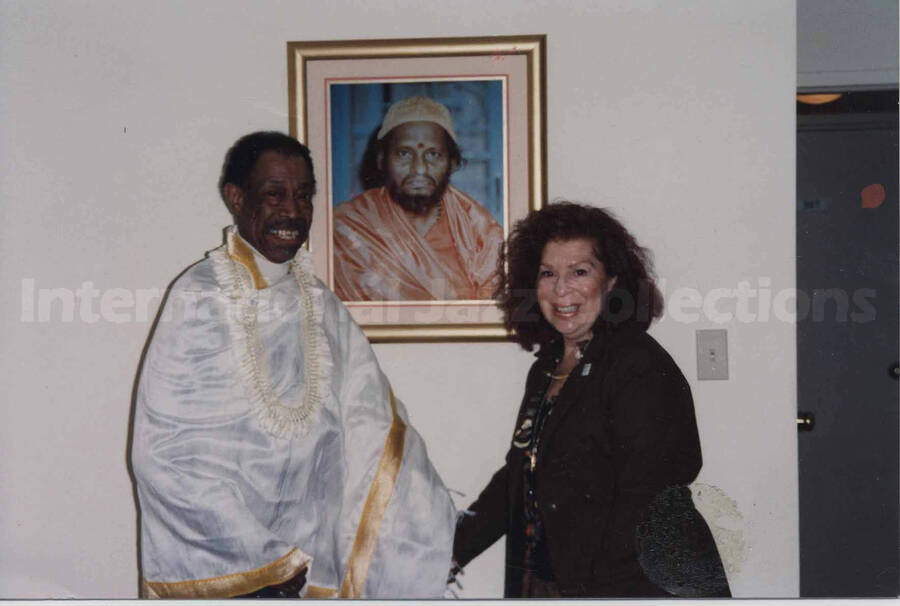 Al Grey wearing what appears to be a ceremonial costume posing with Rosalie in front of a picture of an unidentified man, probably a religious or political leader