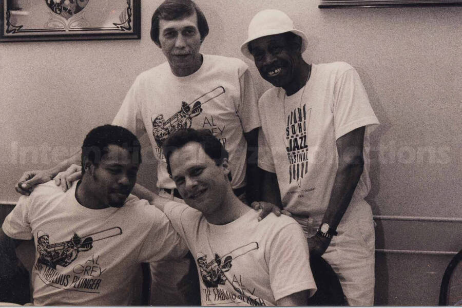 Al Grey posing with three unidentified men. Al Grey's shirt reads: Soho Jazz Festival. The men's shirts read: Al Grey, the Fabulous Plunger