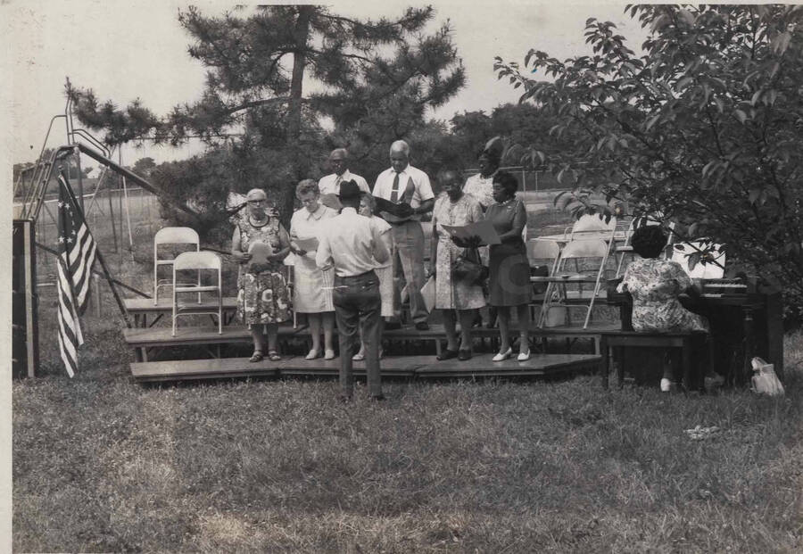 Unidentified persons singing on a stage at a playground included are a pianist and a conductor. A label on the back of the photograph reads: Pennhurst State School and Hospital, Spring City, PA