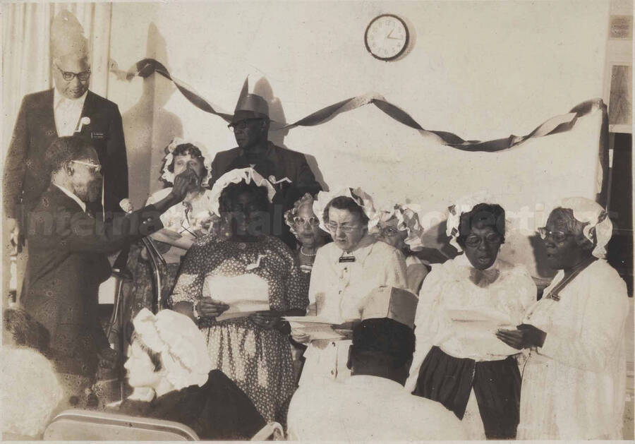 Unidentified women singing, including a conductor, and some of the audience