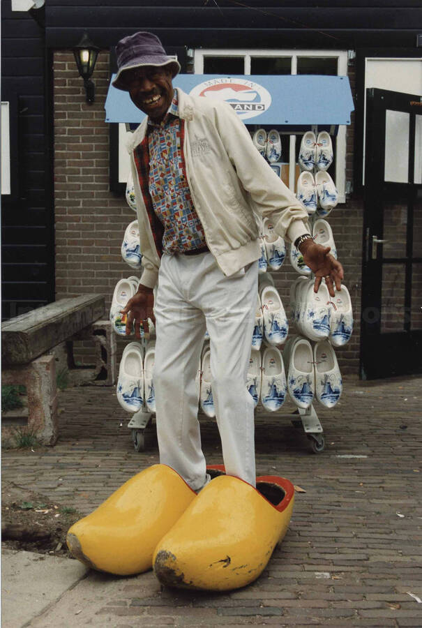 Al Grey trying on oversize traditional wooden shoes clogs from Holland. A label on the back of the photograph reads: Saturday 1991-08-10. 65th Anniversary Michiel de Ruyter (Mike);  island "Marken"/The Netherlands