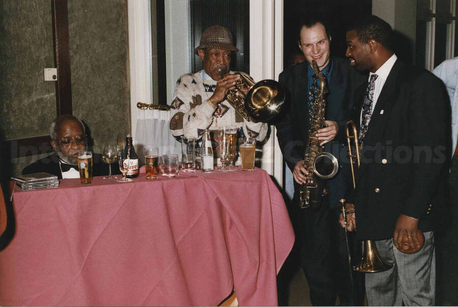 Al Grey playing the trombone alongside two unidentified musicians, one a saxophonist and the other a trombonist next to an unidentified man at a tavern. A label on the back of the photograph reads: Sunday 1992-11-01.  Jazz Mecca 1992/Maastricht-The Netherlands; Golden Tulip/Barbizon Hotel-Maastricht