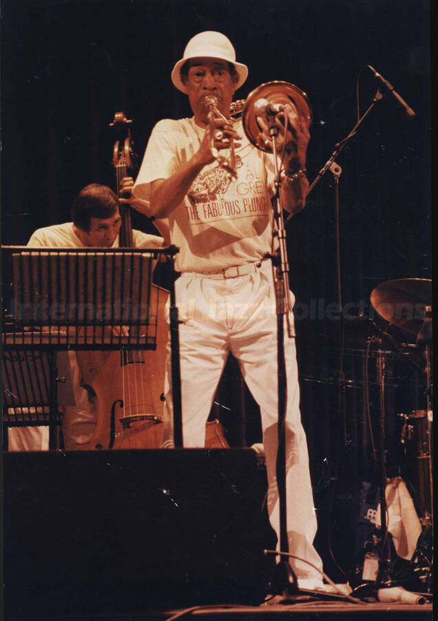 Al Grey performing. Behind him, on the stage, is an unidentified bassist. Al Grey is wearing a shirt that reads: Al Grey, The Fabulous Plunger. A label on the back of the photograph reads: Thursday 1990-07-12; North Sea Jazz Festival 1990/Congresgebouw-The Hague/Rembrantdtzaal