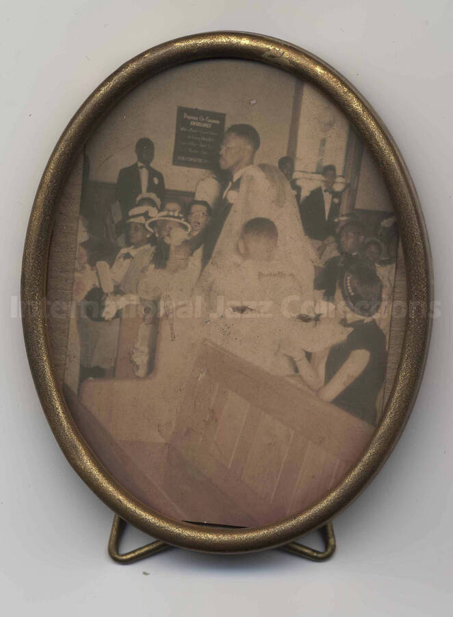 A wedding picture of Richard Grey walking his daughter down the central aisle of a church observed by the guests on both sides. The photograph is in a 3 1/2 x 2 3/4 inch oval metal desk frame