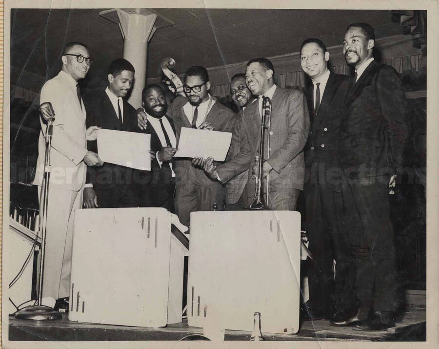 Al Grey posing with seven unidentified men. Two are holding award certificates. This photograph is pasted on a photo album