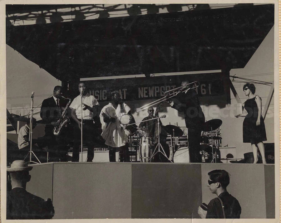 Unidentified musicians performing on a stage. A banner on the back of the stage reads: Music at Newport 1961. This photograph is pasted on a photo album