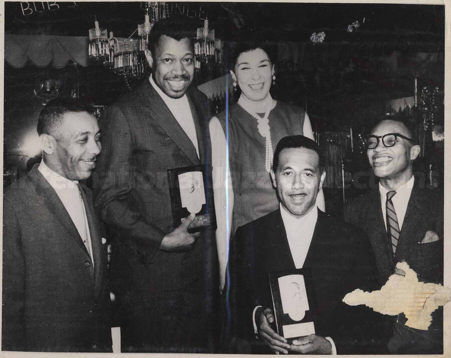 Al Grey posing with Ella Fitzgerald and Count Basie's guys. Al Grey and one of the men are holding award plaques. This photograph is pasted on a photo album