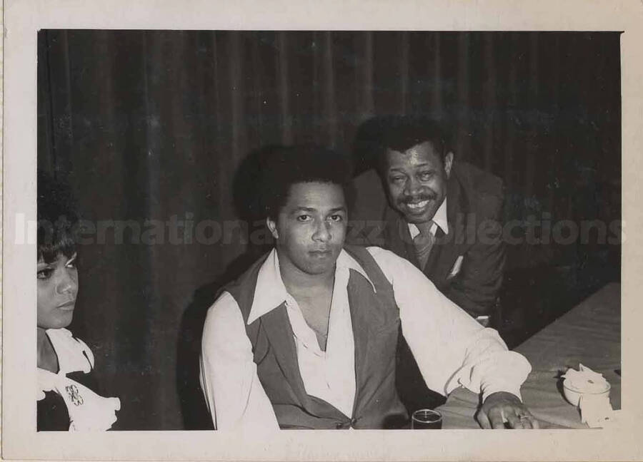 Al Grey posing with unidentified persons at a table in a restaurant. This photograph is pasted on a photo album sheet.