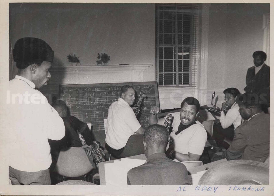 Al Grey and unidentified musicians sitting in front of a blackboard with the scores of the song Round Midnight written on it.with unidentified student, who is playing the guitar, at the Philadelphia Parkway School.;  This photograph is pasted on a photo album sheet.