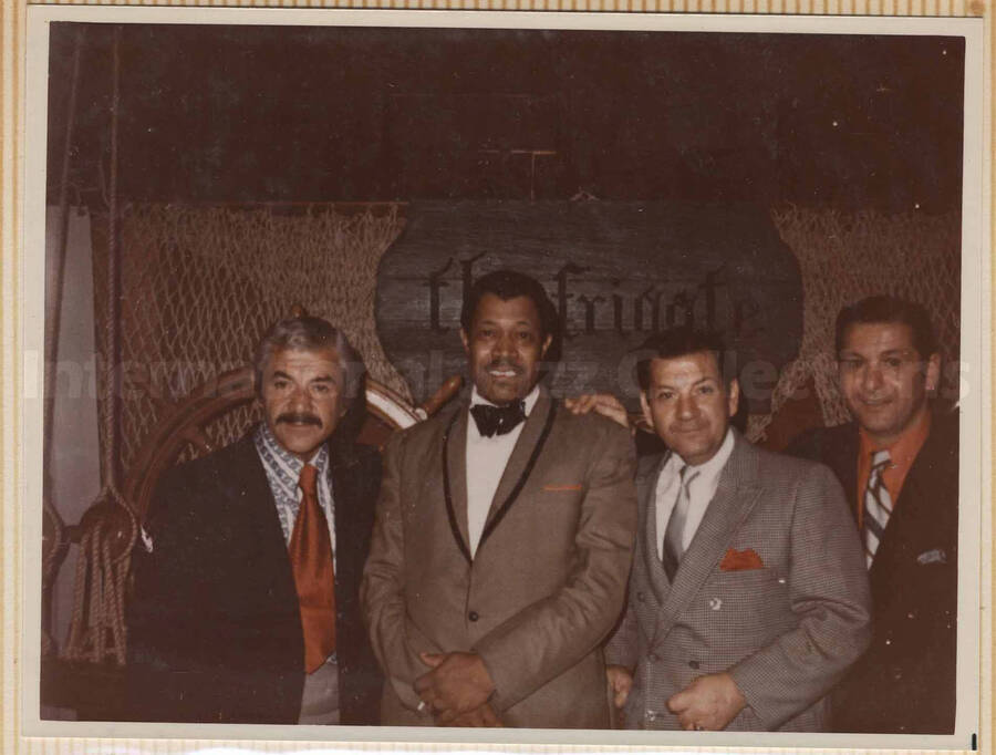 Al Grey posing with three unidentified men. A sign on the back wall reads: The Frigate. This photograph is pasted on a photo album sheet.