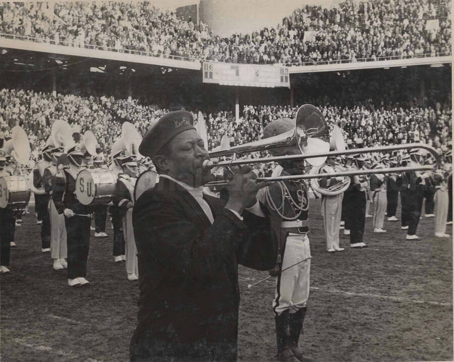 Al Grey playing the trombone in front of an unidentified band in a crowded stadium. The members of the band are wearing uniforms with the letter "S" on their chest and the drums show the initials "S.U.". This photograph is pasted on a photo album