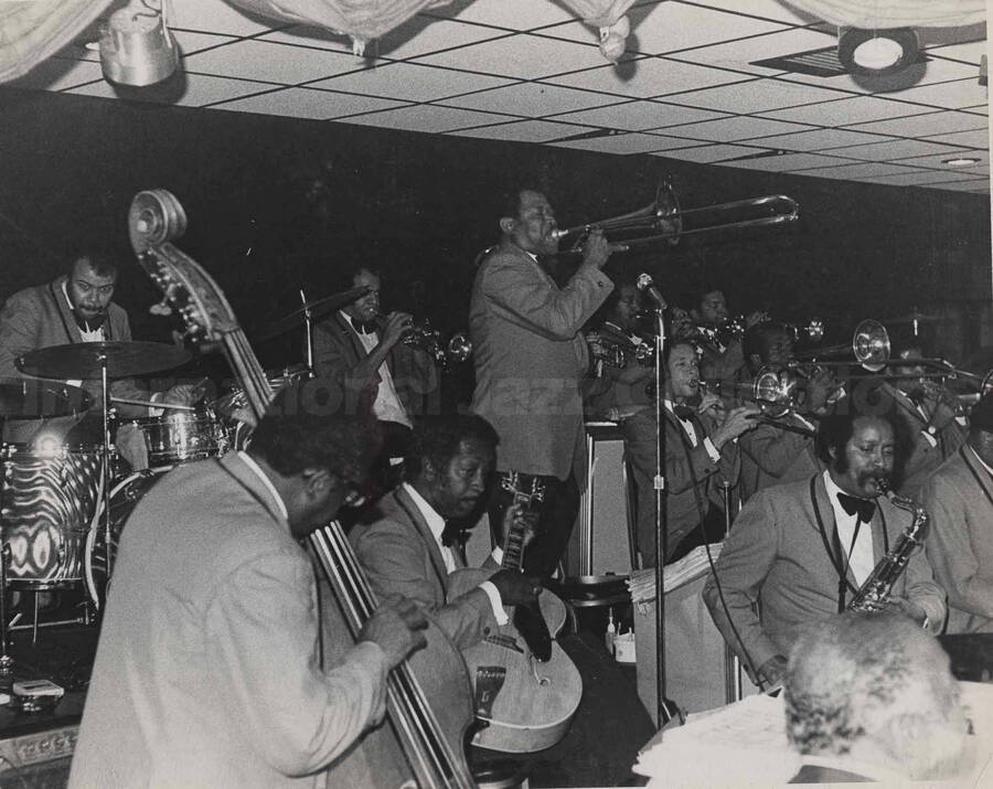 Al Grey performing in an unidentified orchestra featuring also drummer Harold Jones and guitarist [Freddie Green]. This photograph is pasted on a photo album