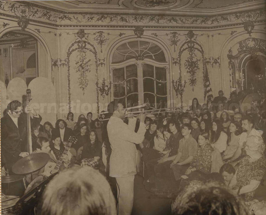 Al Grey and unidentified band performing for an audience comprised mostly of youths in a saloon decorated with musical motifs. This photograph is pasted on a photo album