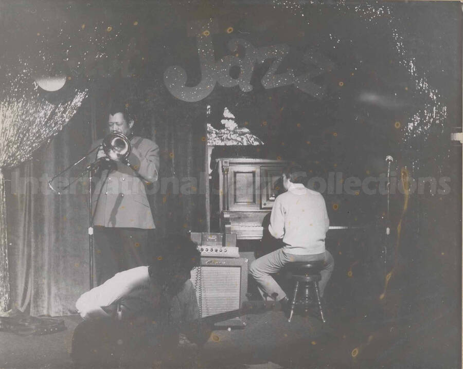 Al Grey performing with two unidentified musicians, one a pianist and the other a guitarist. Written on the background wall: Just Jazz. This photograph is pasted on a photo album