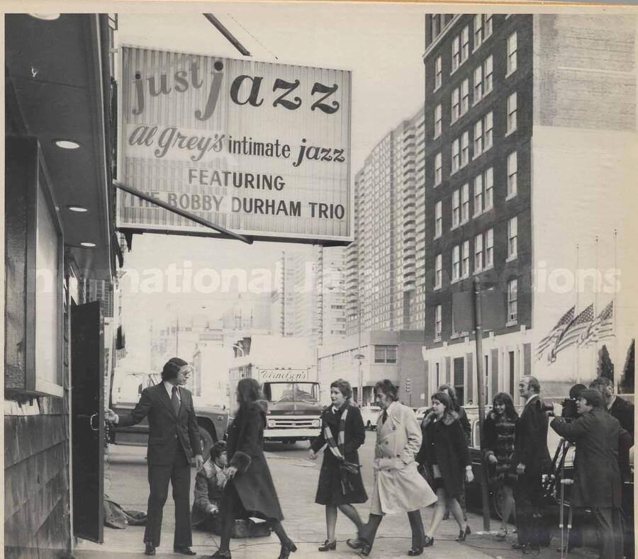 A sign above a door reads: Just Jazz, Al Grey's intimate jazz, featuring the Bobby Durham Trio. An unidentified man holds a door open for the customers, all of this filmed by an unidentified cameraman. On the other side of the street three United States flags are at half mast. This photograph is pasted on a photo album