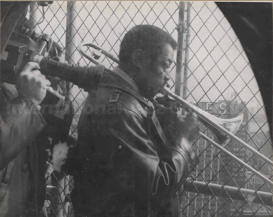 Al Grey playing the trombone on top of a building while the scene is filmed by an unidentified cameraman on his back right side. This photograph is pasted on a photo album