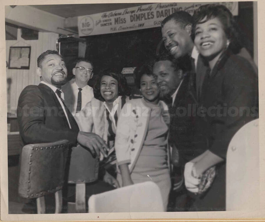 Al Grey posing with three unidentified women and three unidentified men. A banner stretched on the background reads: singer and dancer Miss Dimples Darling. This photograph is pasted on a photo album