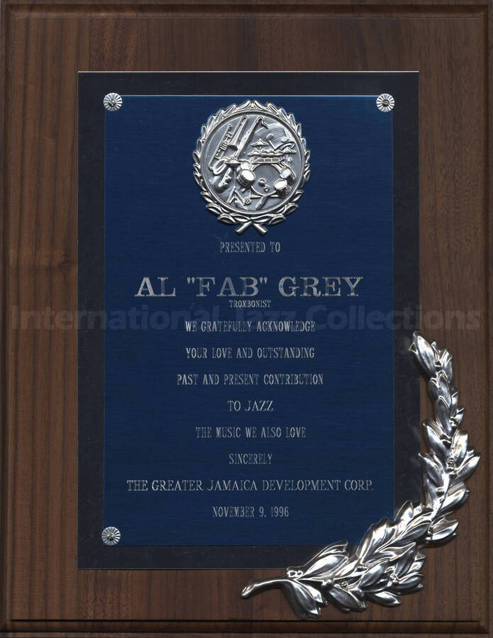 Wood finish plaque with double engraved plate bearing a disk and a laurel leave on the right lower corner. To Al Grey from the Greater Jamaica Development Corporation. A label on the back of the plaque reads: American Roots Art Gallery, St. Albans (NY)