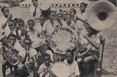 A band composed of boys. Written on the drum: S. D. A. School Osa-Uku. This is a clip from a magazine glued to a piece of cardboard