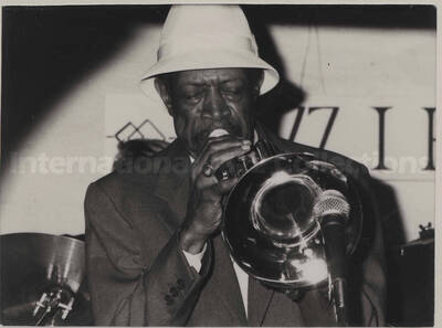 Al Grey playing the trombone. The photograph is pasted on a white mat, which bears a signed dedication to Al Grey for his birthday from Werner Pavei. The photograph is under glass in a 12 x 10 aluminum frame. Illegible handwritten notes on the back of the frame, including the name Monchsberg? [Salzburg, Austria]