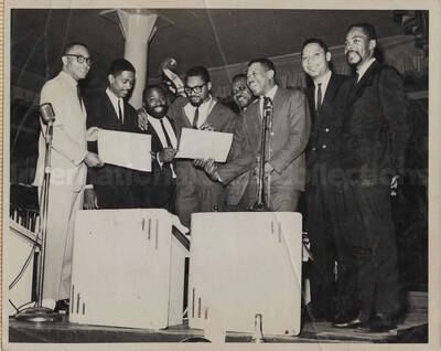 Al Grey posing with seven unidentified men. Two are holding award certificates. This photograph is pasted on a photo album