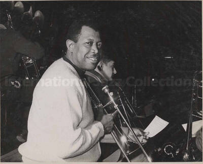 Al Grey holding his trombone and a piece of paper with musical score in the company of unidentified musicians. This photograph is pasted on a photo album
