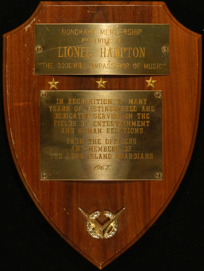Plaque. 12"x9" Shield wood finish plaque with two engraved plates, three small stars between them and a wreath on the bottom of the plaque Honorary Membership presented to Lionel Hampton by the Officers and Members of the Long Island Guardians, in recognition of many years of dedicated service in the fields of entertainment and human relations. 1967