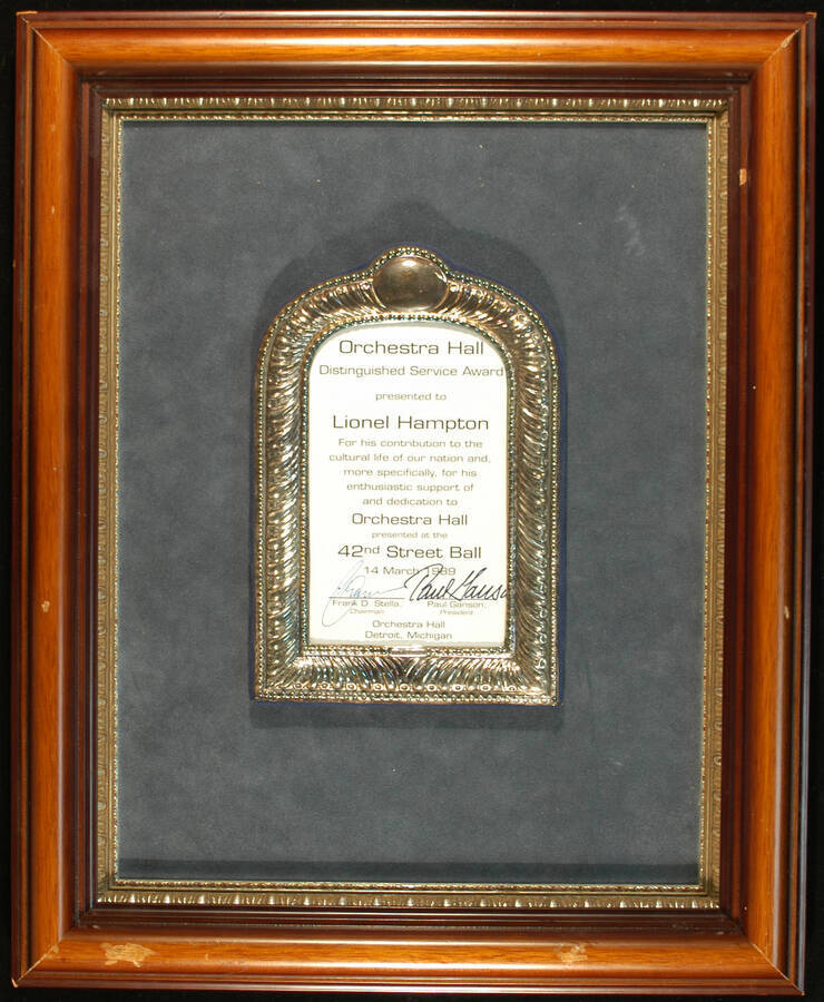 Plaque. 16 1/2"x13 1/2" Wood frame holding a 7 1/2"x5"  silver frame with certificate under glass, in a  and blue velvet background Orchestra Hall Distinguished Service Award presented to Lionel Hampton for his enthusiastic support and dedication to Orchestra Hall, on occasion of the 42nd Street Ball. Frank D. Stella, Chairman and Paul Ganson, President. Detroit, MI, Mar. 14, 1989
