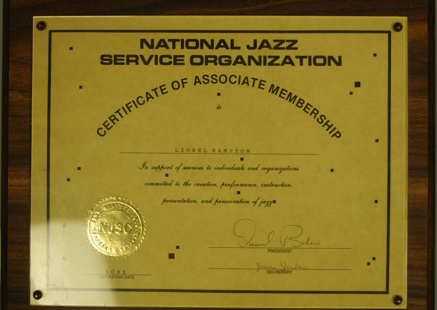 Certificate Plaque. 8 1/2"x11" Certificate with gold foil seal and red ribbon, mounted under acrylic on a 10 1/2"x13" wood plaque Certificate of Associate Membership presented to Lionel Hampton by the National Jazz Service Organization - NJSO in support of services to individuals and organizations committed to the creation, performance, instruction, presentation, and preservation of jazz.