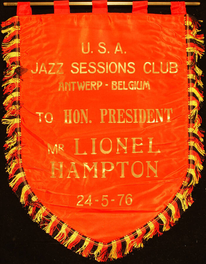 17"x14" Red flag with red, black and yellow fringes around. It bears a printed gold dedication on one side. On a label on the reverse it reads "Antwerp-Belgium. U.S.A. The Best in Swinging Jazz. Jazz Sessions Club. Jazz. Home Place Redout's Gravenwezel." Also a 13" long hanger To Lionel Hampton from the U.S.A. Jazz Sessions Club Antwerp - Belgium. May 24, 1976