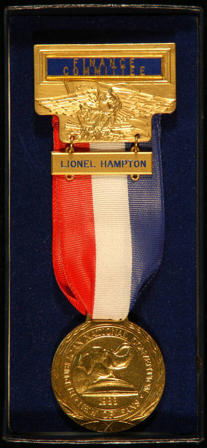 Medal, Pin and Name Badge.  1 1/2" Gold medal with the inscription: "Republican National Convention. 1988. New Orleans." The medal hangs from a red, white and blue ribbon attached to a 1 1/4"x2" gold pin depicting an eagle holding a bundle of arrows with a flag in the background. Above the eagle the pin bears an engraved blue enameled plate that reads "Finance Committee." An engraved plate with Lionel Hampton's name hangs from the pin. This 5"x2" item is inside a blue cardboard presentation box with no cover. A label glued to the back of the box reads "Finance Committee. Badge Code: II: A. BD. Dir/Eagle/Pres. Trust. Lionel Hampton"