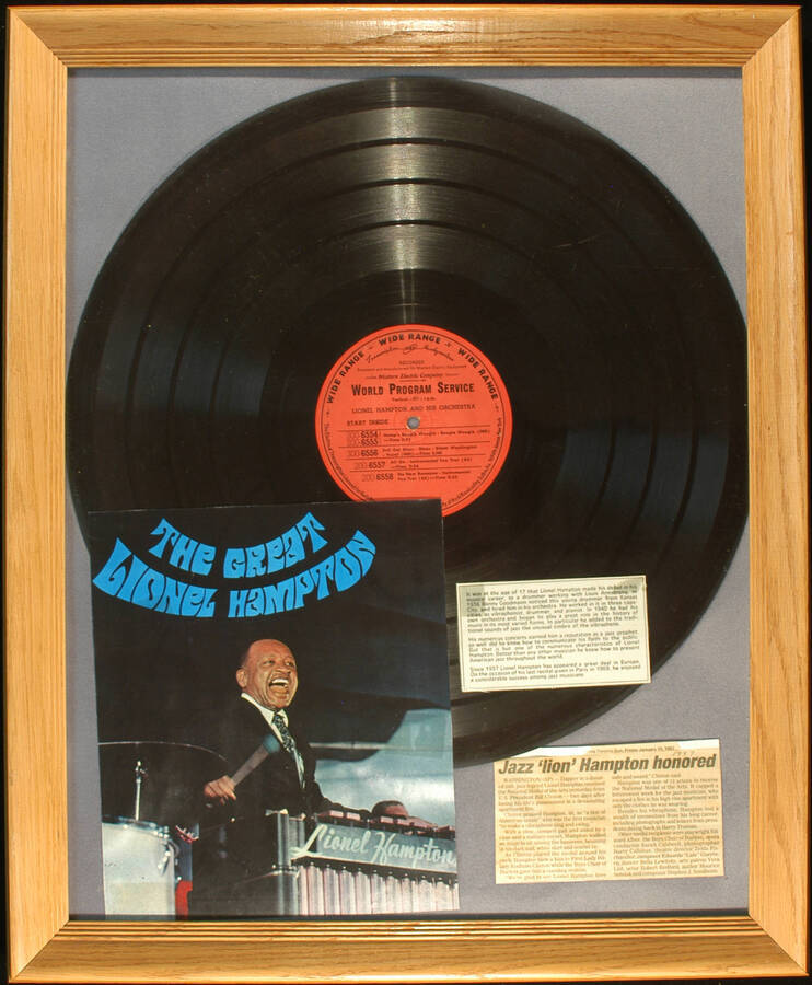 Plaque. 22"x18 1/4" Framed board displaying a 16" acetate record of the "World Program Service" containing the following songs: "Hamp's Boogie Woogie, Boogie Woogie (IND) (200-6554, 200-6555), Evil Gal Blues, Blues, Dinah Washington (IND) (300-6556), All On, Instrumental Fox Trot (AS) (200-6557), No New Romance, Instrumental Fox Trot (AS) (200-6558), a color picture of Lionel Hampton, a short biography, and a clipping from The Toronto Sun about receiving the National Medal of Arts from U.S. President Bill Clinton, two days after his apartment got on fire.