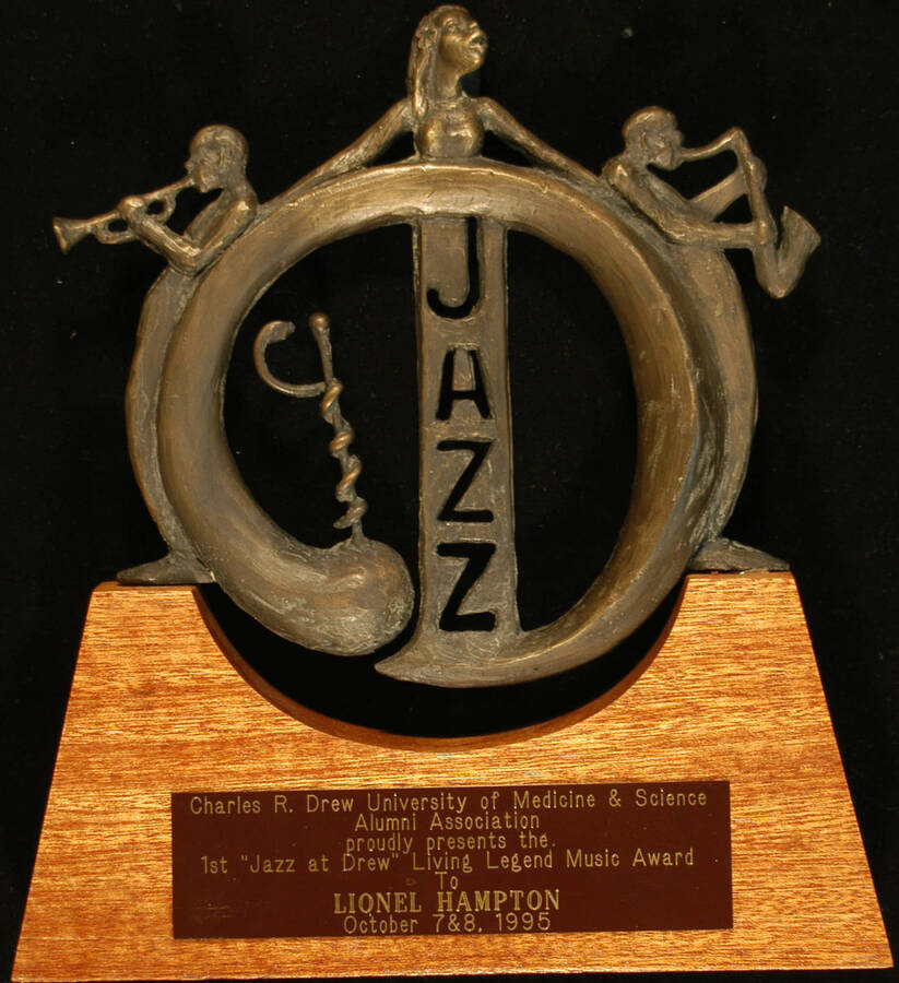 Trophy.  9x10 Circular bronze sculpture depicting, inside, the word “jazz” and the rod of Asclepius, consisting of a serpent entwined around a staff.  Outside, it is flanked by two musicians playing trumpet and saxophone, and on top, a bust of a female singer. It is mounted on a wood base with engraved plate and rests on an oblong flat piece of stone. On the back, it bears the artist's autograph and a date "Charles Dickson" "5-16-95" traced into the bronze First "Jazz at Drew" Living Legend Music Award presented to Lionel Hampton by Charles R. Drew University of Medicine and Science Alumni Association. Los Angeles, CA, Oct. 7-8, 1995